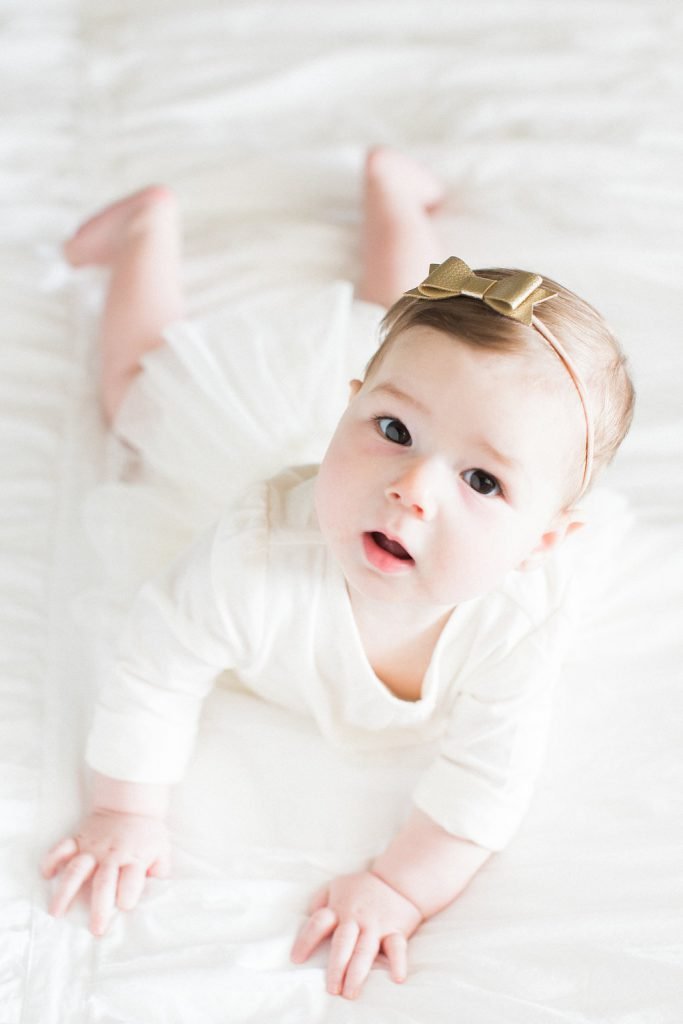 Photo of baby lying on a bed in a white dress with a gold bow in her hair