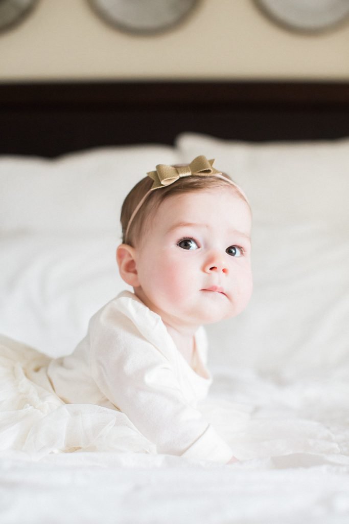 Picture of baby on a bed with white sheets in a white dress with a gold bow