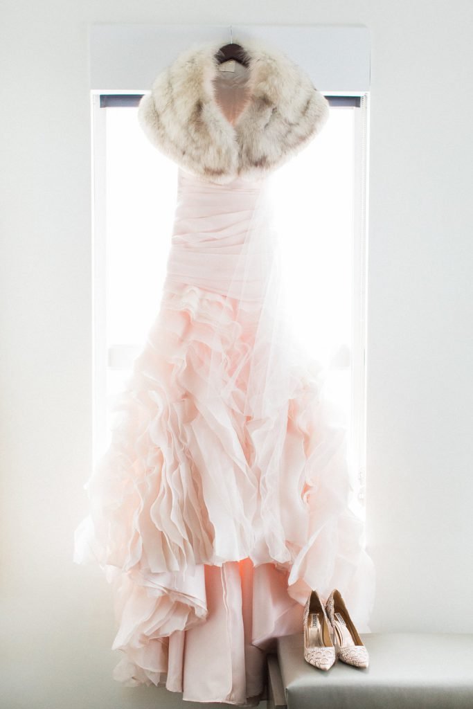 Photograph of blush pink wedding dress hanging in hotel room with fur shawl and badgley mischka bridal shoes