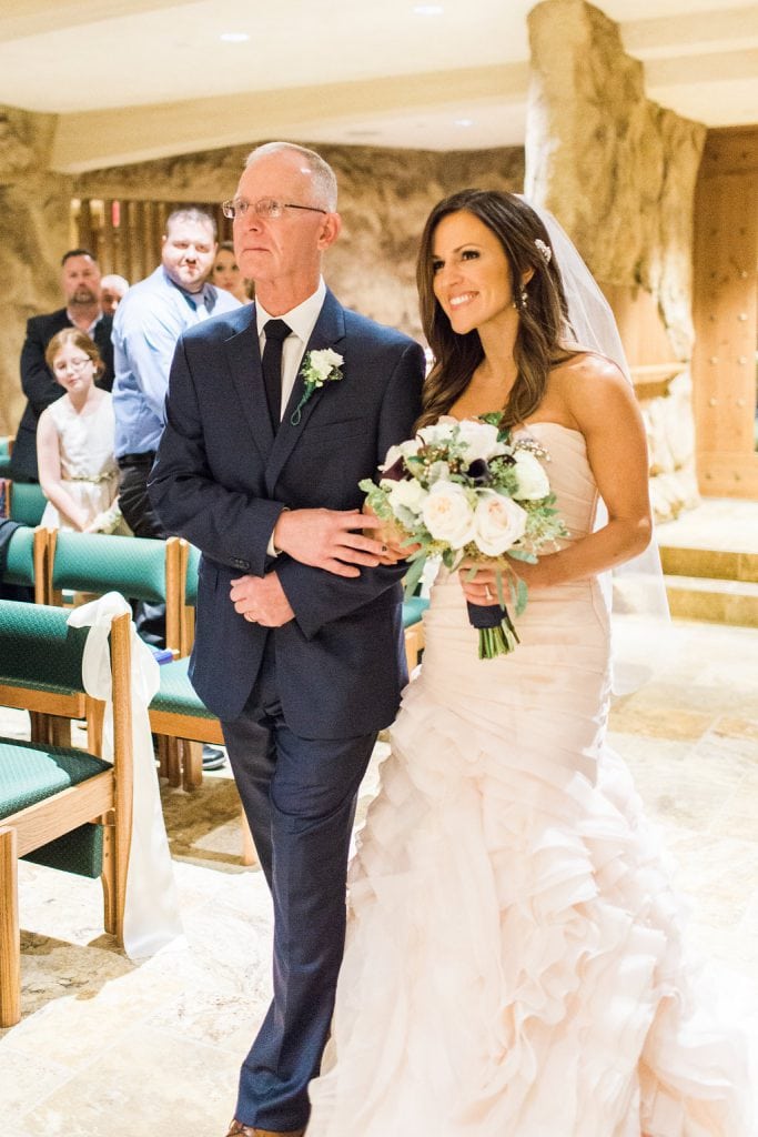 The bride being walked down the aisle by her father in the grotto of the Saints John and Paul Roman Catholic Church