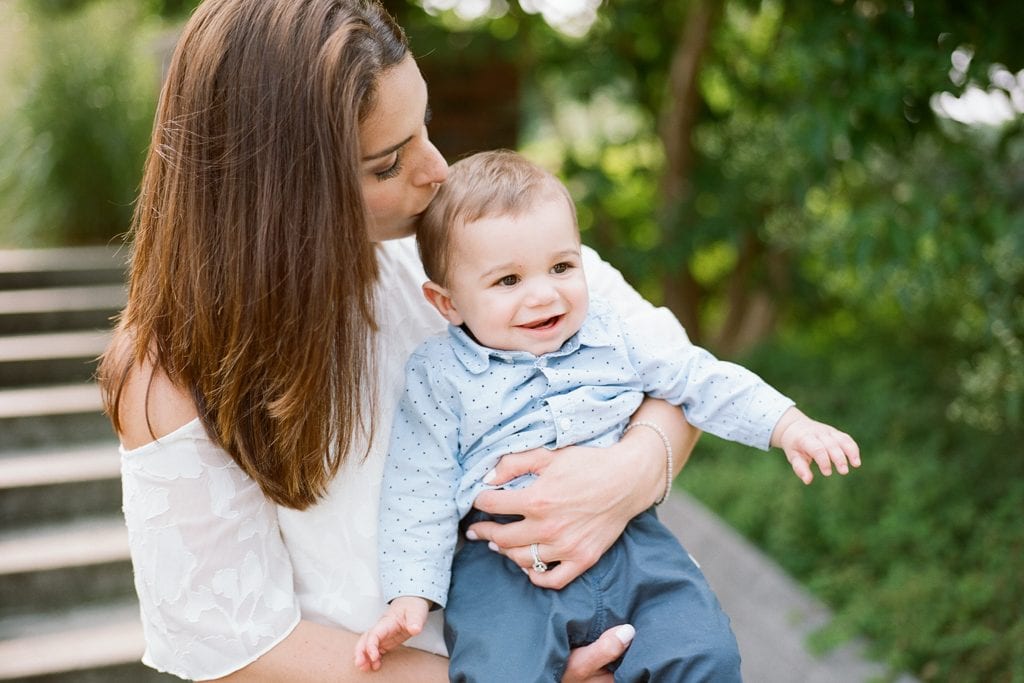 Mom kissing baby son on the head at the park wearing a white off the shoulder top during Family Session at The Walled Garden