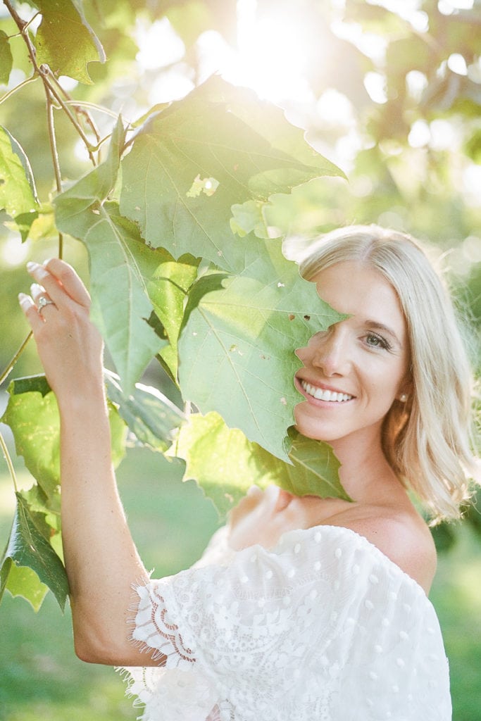 Beautiful blue eyes and blonde hair woman standing under a tree with leaves around her wearing white ruffled show me your mumu dress