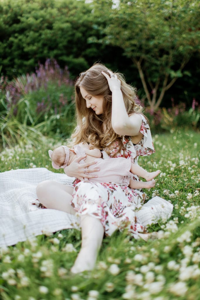 Young mother wearing a light pink floral dress breastfeeds her newborn baby girl while sitting in the grass: outdoor newborn session