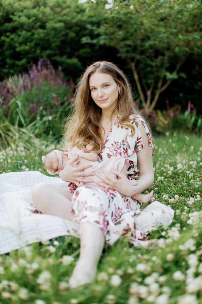 Young mother wearing a light pink floral dress breastfeeds her newborn baby girl while sitting in the grass: outdoor newborn session