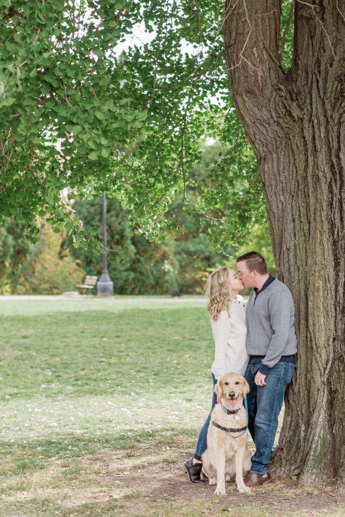 Couple kissing against a tree holding their dog