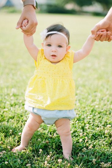 Little girl holding her parents hands while standing in grass wearing a yellow top