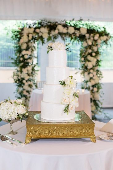 white wedding cake with white flowers in front of a white flower arch