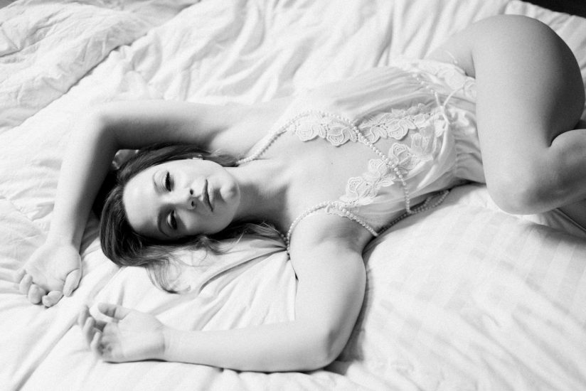 woman laying on a bed in a white lace romper with pearls around her neck: Pittsburgh Boudoir Photography